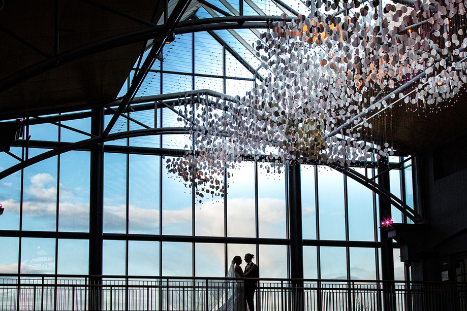 Bride and groom stand in front of large windows with beautiful sky and light fixture